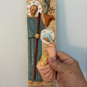 May include: A rectangular, beige relief sculpture depicting a religious scene. It features figures of Joseph, Mary, and baby Jesus, along with a donkey and a calf. The figures are in blue, pink, and yellow. The artist's signature is at the bottom.