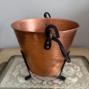 May include: A hammered copper bucket with a black metal handle and stand. The bucket has a flared shape and a shiny, reflective surface. The black metal handle is attached with rivets. The bucket is a decorative item.