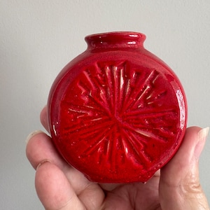 May include: A small, round, red ceramic vase with a textured, starburst design.