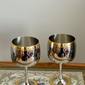 May include: Two stainless steel wine glasses with gold rims and bases. The glasses have a reflective surface, and the gold accents add a touch of elegance. The stem is slender, and the base is circular.