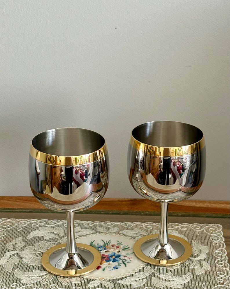 May include: Two stainless steel wine glasses with gold rims and bases. The glasses have a reflective surface, and the gold accents add a touch of elegance. The stem is slender, and the base is circular.
