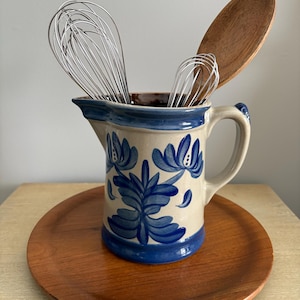 May include: A blue and white ceramic jug with floral designs, holding two wire whisks and a wooden spoon. The jug is sitting on a wooden tray.