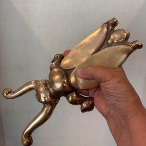May include: A polished brass sculpture shaped like a large insect with prominent wings and curved legs. The metallic object features a smooth, reflective surface with a golden hue, showcasing detailed segments on the body and elongated, curled limbs.
