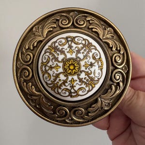 Peut inclure: Un bouton de porte vintage en laiton avec un centre en céramique florale blanche et jaune. Le bouton a un design orné et tourbillonnant sur le bord. Le centre présente un motif floral jaune sur fond blanc.