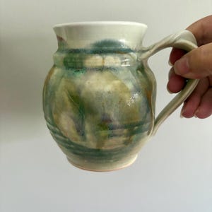 May include: A ceramic mug with a rounded body and a flared rim, featuring a glossy glaze in blended shades of sage green, earthy brown, and cream. The mug has a curved handle on the side and horizontal ridges around the lower section of the base.