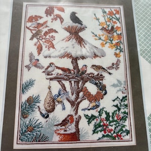 May include: A cross-stitch pattern depicting a bird feeder with a variety of birds, including robins, bluebirds, cardinals, and sparrows. The feeder is surrounded by greenery, including holly and pine branches.