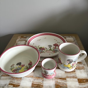 May include: A white ceramic tableware set with a pink trim, decorated with illustrations of children and mushrooms. The set includes a bowl, plate, mug, and small cup. The items are displayed on a patterned surface.