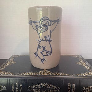 May include: A cylindrical beige ceramic tumbler featuring a dark blue line drawing of a scarecrow with a hat, scarf, and patches. The tumbler is positioned on a black book with gold decorative accents and the text Great Expectations Charles Dickens.