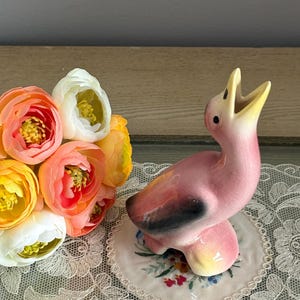 May include: A pink ceramic bird ornament with a yellow beak and black detailing. The bird sits on a floral-patterned base. A bouquet of orange, yellow, and white flowers is to the left. The ornament is on a lace doily.