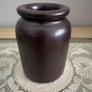 May include: A dark brown ceramic vase with a wide, rounded rim. The vase has a cylindrical body and sits on a white lace doily. The vase is approximately 6 inches tall.