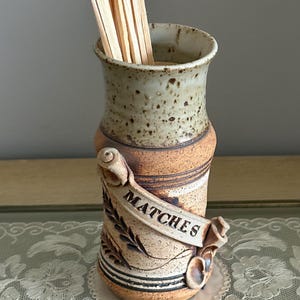 May include: A ceramic match holder with a speckled, light brown and tan finish. The word "MATCHES" is displayed on a banner wrapped around the holder. The holder contains wooden matches.