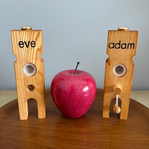 May include: Two wooden stands with the words "eve" and "adam" inscribed on them. A red apple sits between the stands.