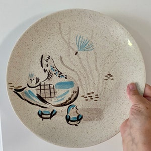 May include: A white ceramic dinner plate with a speckled glaze features a stylized design of a brown and blue bird with two chicks. The design is in a mid-century modern style.