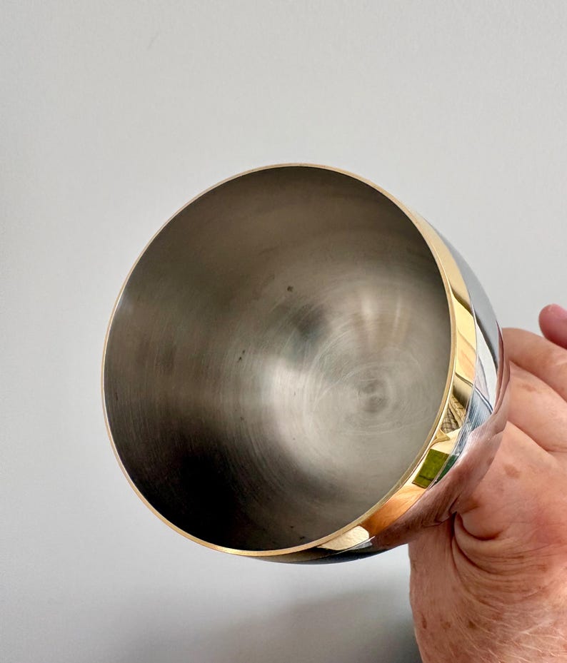 May include: A stainless steel cocktail shaker with a gold band around the top. The interior of the shaker is a brushed metal finish. The shaker is held by a person's hand against a plain white background.
