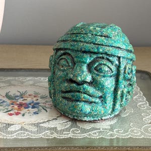 May include: A teal-colored, textured sculpture of a human head with detailed facial features and a headdress. The sculpture is placed on a decorative tray with a floral design.