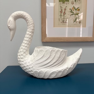 May include: A white swan-shaped planter with a textured finish. The swan features a curved neck and detailed feather patterns. It rests on a dark blue surface, with a framed botanical print visible in the background.