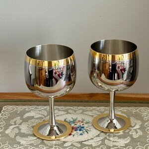 May include: Two stainless steel wine glasses with gold rims and bases. The glasses have a reflective surface, and the gold accents add a touch of elegance. The stem is slender, and the base is circular.