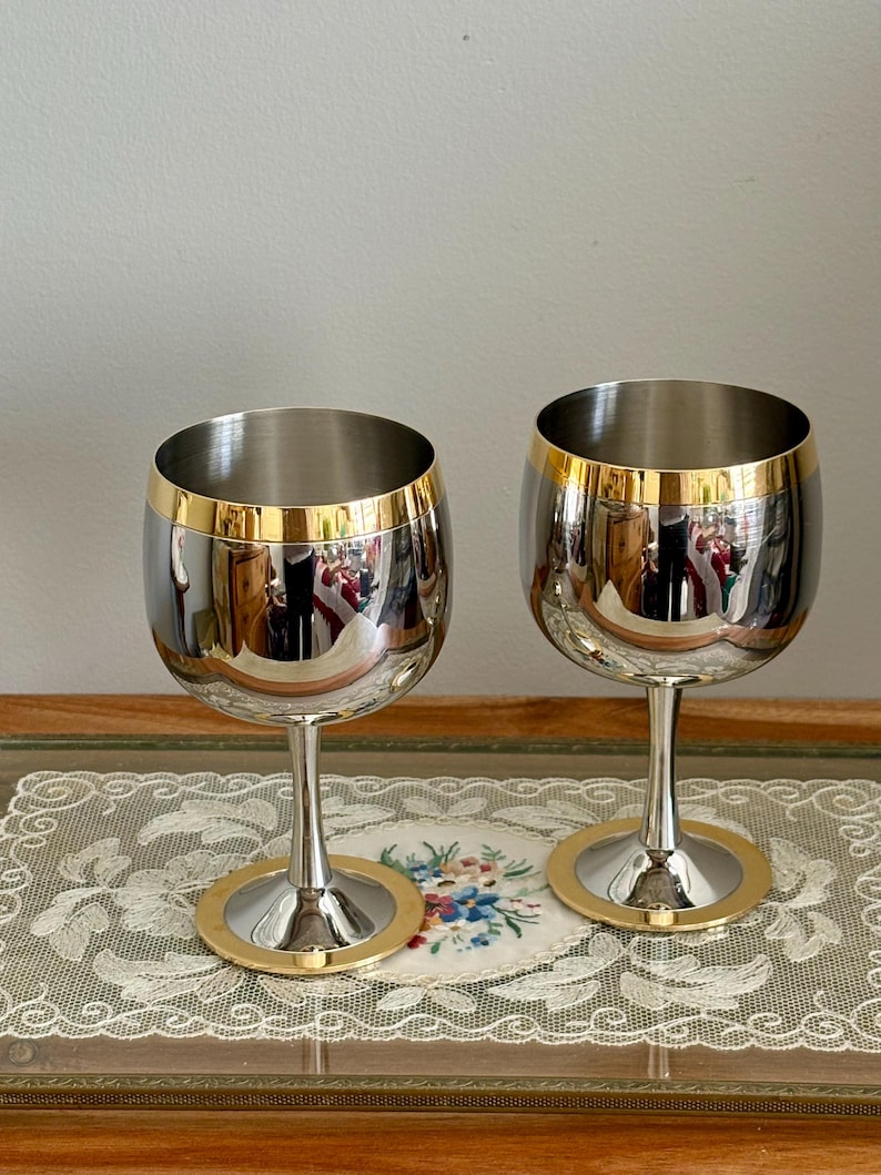 May include: Two stainless steel wine glasses with gold rims and bases. The glasses have a reflective surface, and the gold accents add a touch of elegance. The stem is slender, and the base is circular.