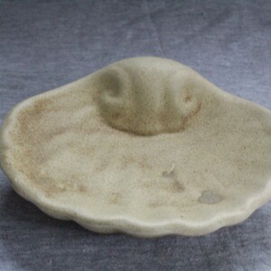 May include: A small, off-white ceramic dish shaped like a seashell with a raised, rounded centre. The dish has a textured surface and a slightly irregular shape.