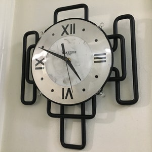 May include: A black metal clock with Roman numeral markings on a white face. The clock is mounted on a white wall.