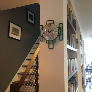 Puede incluir: Un reloj verde y blanco con números romanos en una pared blanca. El reloj tiene la forma de un cuadrado con esquinas redondeadas. El reloj está colgado en una pared blanca junto a una estantería llena de libros.
