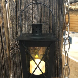 May include: A black metal lantern with a glass panel and a candle inside. The lantern is hanging from a hook on a piece of weathered wood.