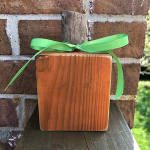 May include: A wooden pumpkin with a rustic brown finish and a green ribbon tied around the top. The pumpkin is sitting on a wooden surface.