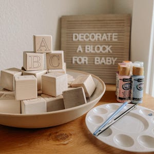 May include: A collection of wooden alphabet blocks, some displaying letters A, B, and C, are arranged in a beige bowl. The blocks are accompanied by paint bottles, a palette, and paintbrushes. A sign reads "DECORATE A BLOCK FOR BABY."