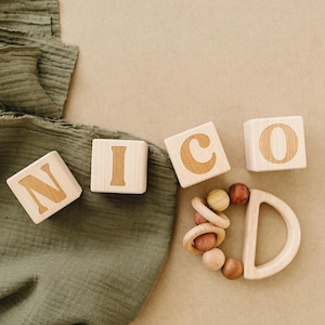 May include: Wooden blocks spelling "NICO" with a wooden baby rattle. The blocks and rattle are light-colored wood with a natural finish. A green fabric is draped on the left side of the image, with a neutral background.