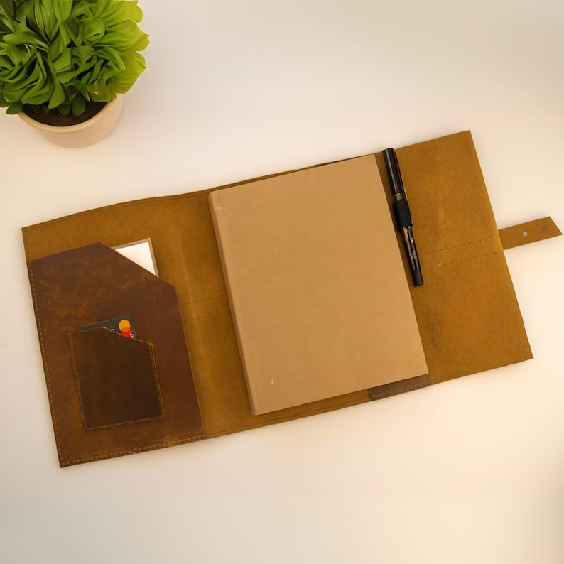 May include: Open, tan leather notebook cover with a brown interior pocket, a notepad, and a black pen. The cover has a tab closure. A small potted plant is in the upper left corner.
