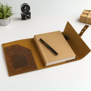 May include: A tan leather journal cover with a brown pocket and a matching strap. A closed, tan-colored notebook and a black and gold pen rest inside the open cover. The journal is on a white surface.