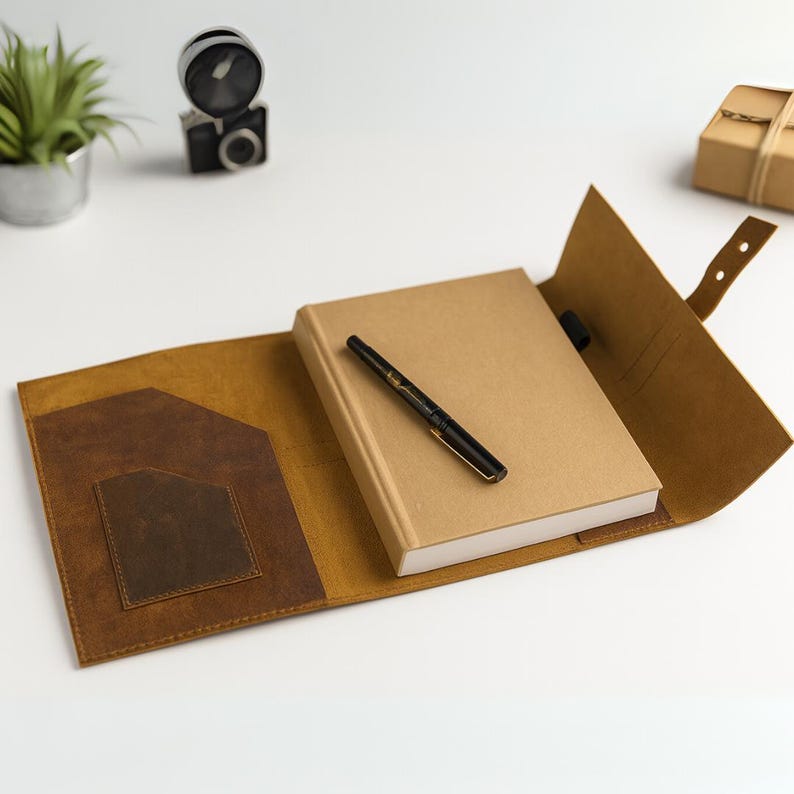 May include: A tan leather journal cover with a brown pocket and a matching strap. A closed, tan-colored notebook and a black and gold pen rest inside the open cover. The journal is on a white surface.