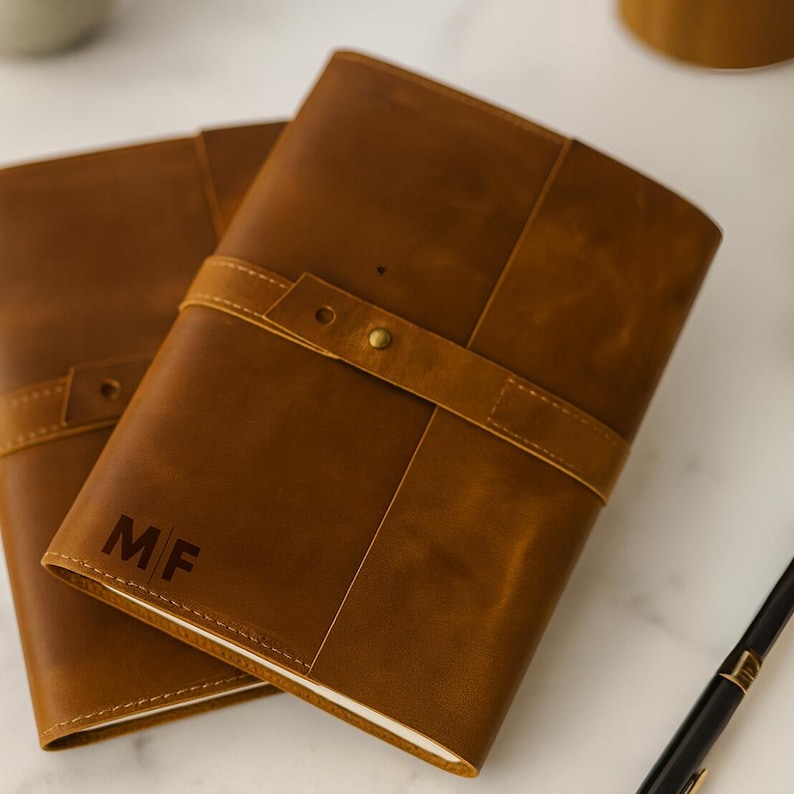 May include: Two brown leather journals with a strap closure and brass hardware. The initials "MF" are embossed on the front cover of one journal. The journals are rectangular and have a classic, elegant design.