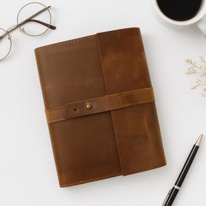 May include: A brown leather journal with a strap closure and brass hardware. The journal is rectangular and appears to be handmade. The image also includes a pen, eyeglasses, and a cup of coffee.
