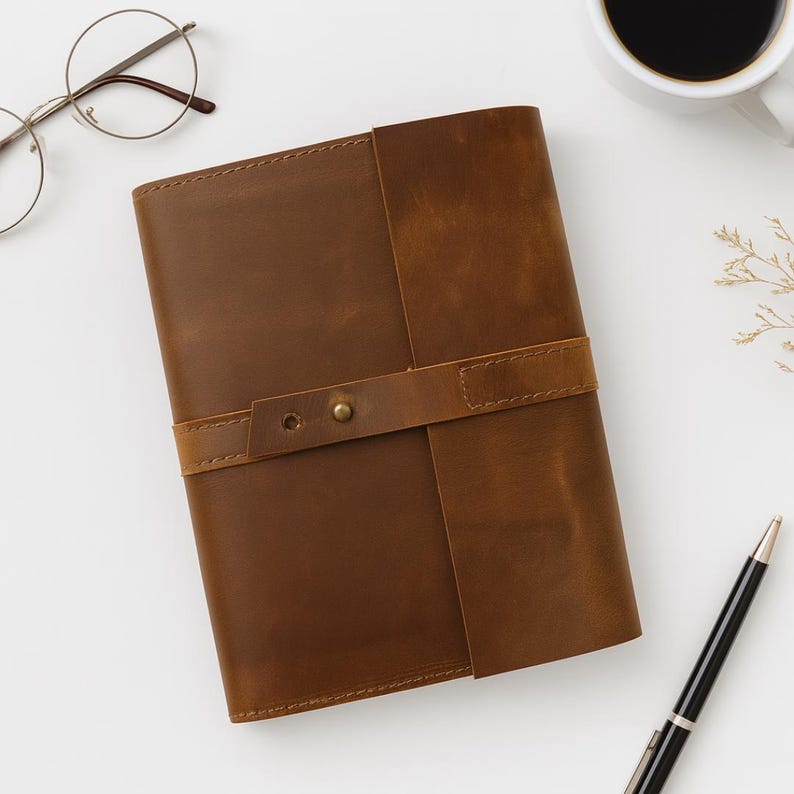 May include: A brown leather journal with a strap closure and brass hardware. The journal is rectangular and appears to be handmade. The image also includes a pen, eyeglasses, and a cup of coffee.