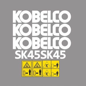 Op de afbeelding: Grijze achtergrond met de witte tekst "KOBELCO" driemaal herhaald, gevolgd door "SK45SK45". Daaronder staan vier gele waarschuwingsborden met zwarte symbolen en figuren.