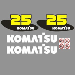 Op de afbeelding: De afbeelding toont twee gele cijfers "25" boven het woord "KOMATSU" in zwarte tekst. Daaronder wordt het woord "KOMATSU" twee keer in het wit herhaald. Kleine rode en witte veiligheidssymbolen zijn ook aanwezig. De achtergrond is grijs.