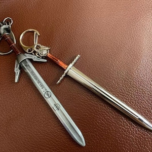 Peut inclure: Deux porte-clés en forme d'épée argentés sur un fond en cuir brun. Le plus grand porte-clés a les mots "Valyrian Steel" gravés sur la lame. Le plus petit porte-clés a un anneau avec un motif décoratif.