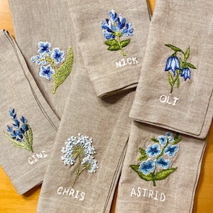 May include: Five linen napkins with embroidered flowers and names. The names are "Cini", "Chris", "Astrid", "Nick", and "Oli".