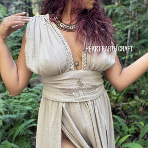 May include: A woman wearing a beige linen dress with a deep V-neckline and a decorative belt featuring an embroidered winged figure. The dress has a slit in the front and the woman is standing in a forest setting. The text "HEART EARTH CRAFT" is visible on the dress.