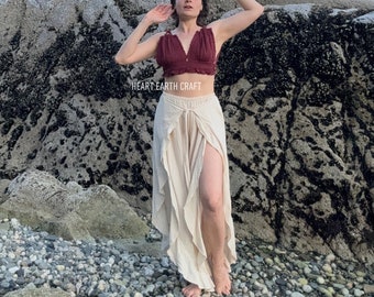 May include: A woman wearing a maroon crop top with a ruffled neckline and a pair of beige, flowy pants with a slit up the side. The pants have a gathered waist and a ruffled hem. The woman is standing on a rocky beach with a blue sky in the background. The text "HEART EARTH CRAFT" is visible on the image.