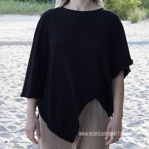 May include: A black cotton poncho with a frayed edge, worn by a person with long blonde hair. The poncho is draped over a brown skirt. The person is standing on a sandy beach.