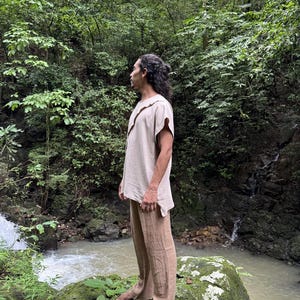 May include: A person wearing a beige linen shirt with a unique collar and matching brown linen pants. The person is standing on a moss-covered rock near a flowing stream, surrounded by lush green foliage.