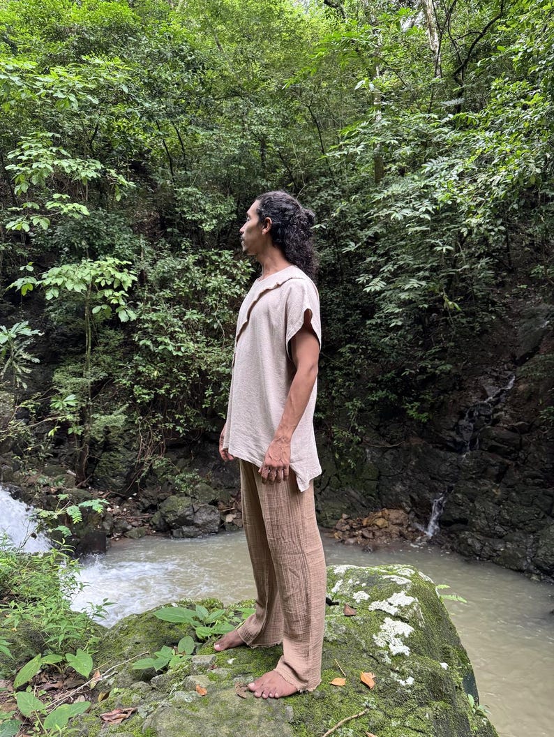 May include: A person wearing a beige linen shirt with a unique collar and matching brown linen pants. The person is standing on a moss-covered rock near a flowing stream, surrounded by lush green foliage.