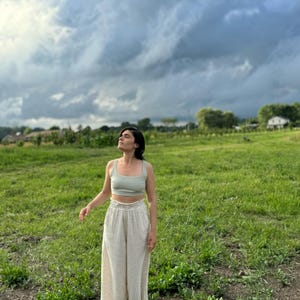 K&ouml;nnte beinhalten: Eine Person tr&auml;gt ein hellgr&uuml;nes Tanktop und eine wei&szlig;e Leinenhose und steht auf einem Feld mit gr&uuml;nem Gras und einem bew&ouml;lkten Himmel.