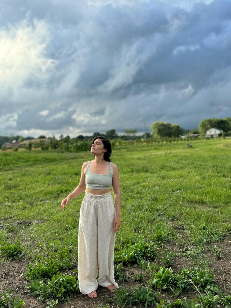 K&ouml;nnte beinhalten: Eine Person tr&auml;gt ein hellgr&uuml;nes Tanktop und eine wei&szlig;e Leinenhose und steht auf einem Feld mit gr&uuml;nem Gras und einem bew&ouml;lkten Himmel.