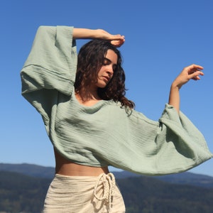May include: A woman wearing a light green, loose-fitting,  cropped top with long sleeves and a cream-colored wrap skirt. The top has a relaxed, flowy silhouette. The skirt is tied at the waist with a long, knotted cord.