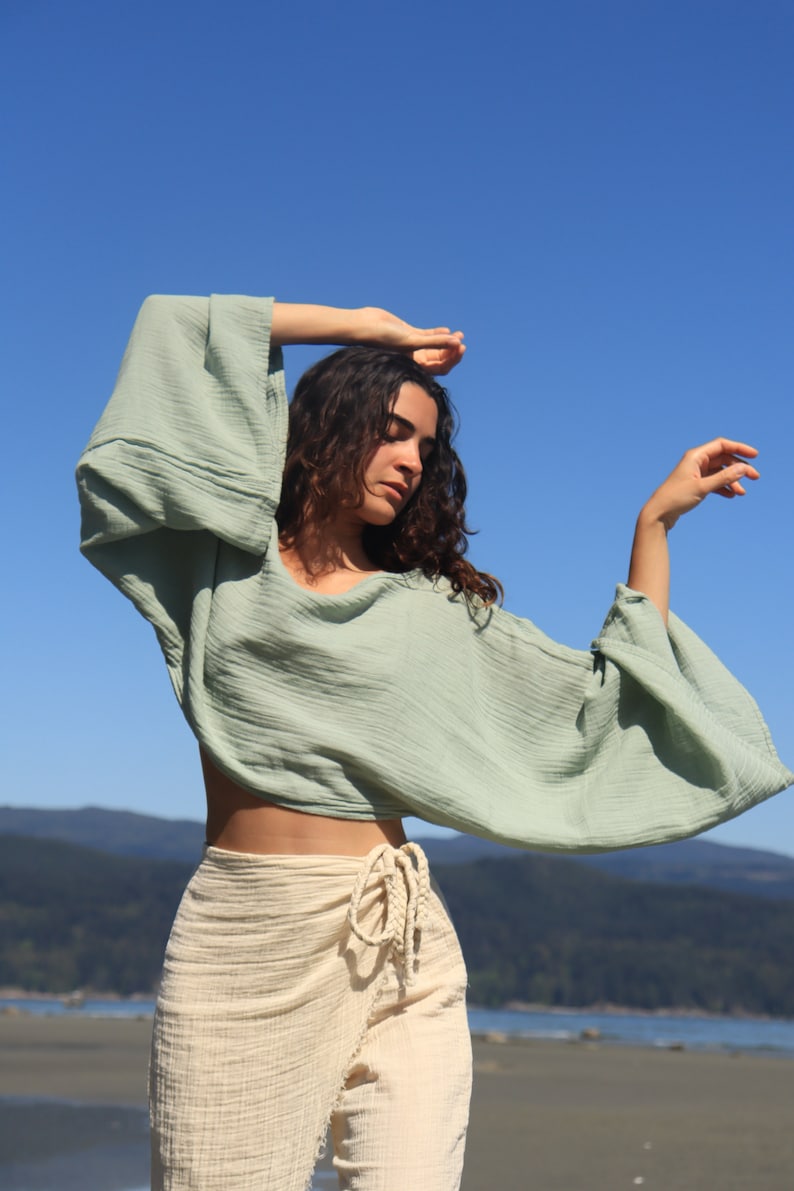 May include: A woman wearing a light green, loose-fitting,  cropped top with long sleeves and a cream-colored wrap skirt. The top has a relaxed, flowy silhouette. The skirt is tied at the waist with a long, knotted cord.