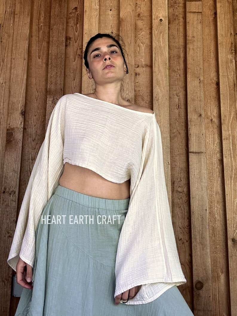 May include: A white, off-the-shoulder, cropped top with long, wide sleeves. The top is made of a lightweight, gauzy fabric. The model is wearing a light blue skirt with a ruffled hem. The text "HEART EARTH CRAFT" is visible on the skirt.
