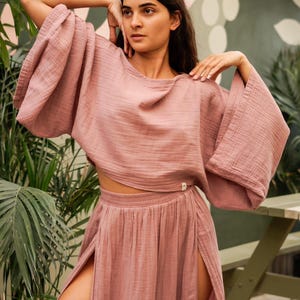 May include: A dusty rose colored two-piece outfit with a cropped top and a long skirt with side slits. The top has wide, flowing sleeves. The skirt has a high waist and a flowy silhouette.